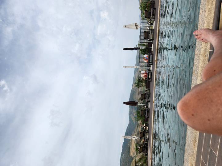 View over a serene infinity pool with loungers and distant hills, foreground shows traveller’s legs