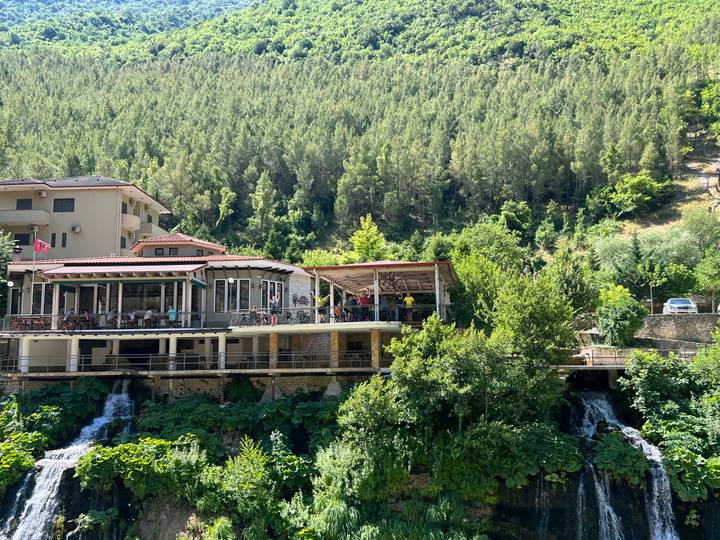 Hillside restaurant complex overlooks a lush valley with small cascades and dense pine forest.