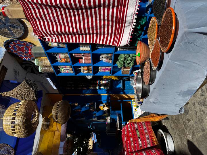 Colorful market stall displays spices, pottery and woven baskets in bright sunlight.
