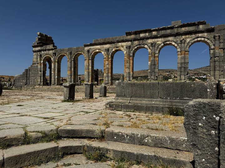 Ancient Roman stone arches stand amid ruined columns under a clear blue Moroccan sky.