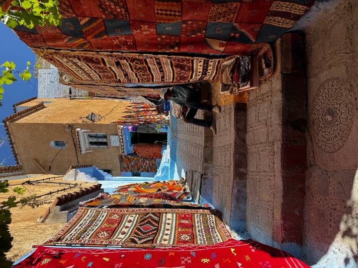 Narrow lane draped with richly patterned carpets leading toward a blue-painted doorway.