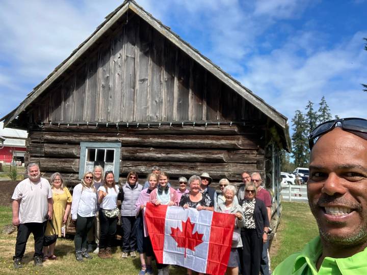 Group of travellers posing with Canadian flag in front of rustic wooden cabin on sunny day