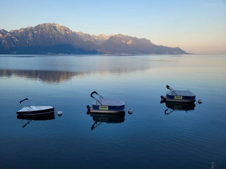Three small motorboats floating on a calm lake reflecting distant alpine peaks at dawn.