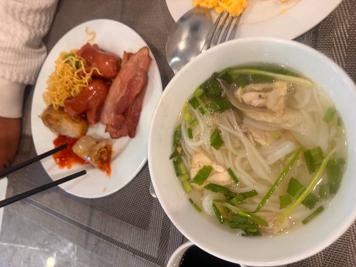 Bowl of steaming pho served with herbs beside a plate of noodles, sausage and spring rolls.