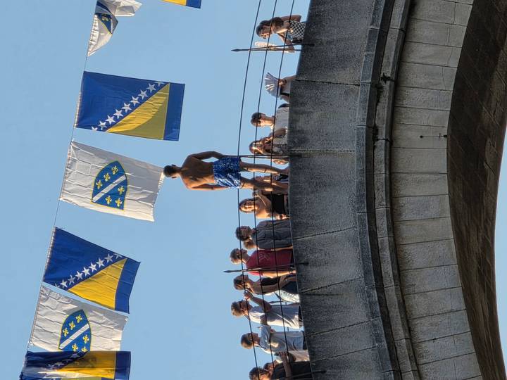 Young man preparing to dive off Mostar’s historic bridge while onlookers gather behind him, Bosnian flags flutter overhead.