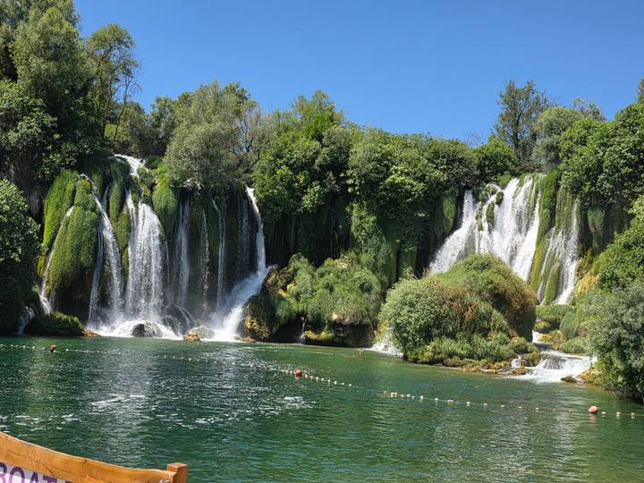 Lush Kravice waterfalls cascading into a clear emerald pool where swimmers enjoy the refreshing water.