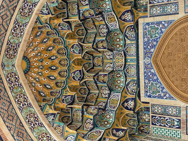 Intricate Islamic tilework ceiling inside a historic madrasah with rich blues, yellows and geometric patterns.