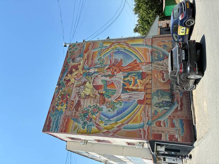 Large colourful mosaic mural on urban building with parked cars below.