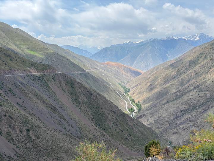 Sweeping high-altitude valley with winding road and distant snow-capped peaks under mixed clouds.