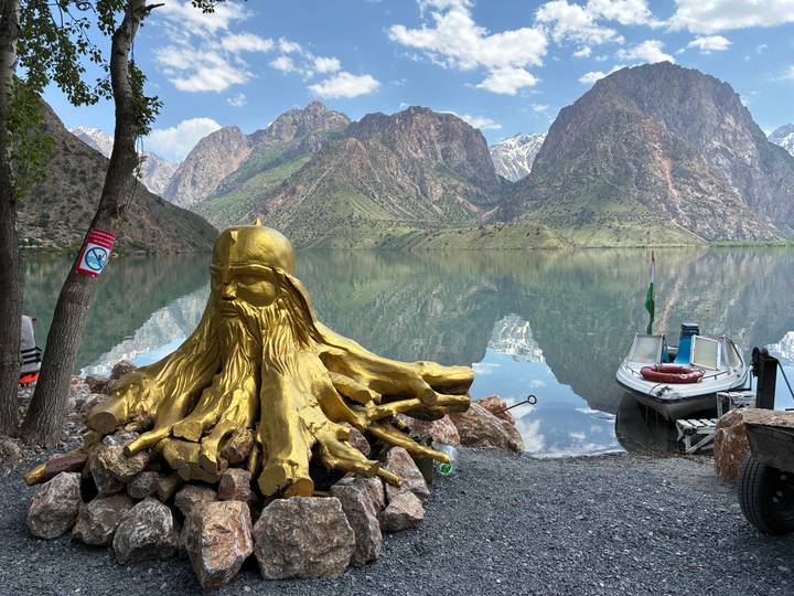 Golden-painted tree-root sculpture beside a tranquil mountain lake with steep green peaks reflecting in the clear water; a small docked boat sits to the right.
