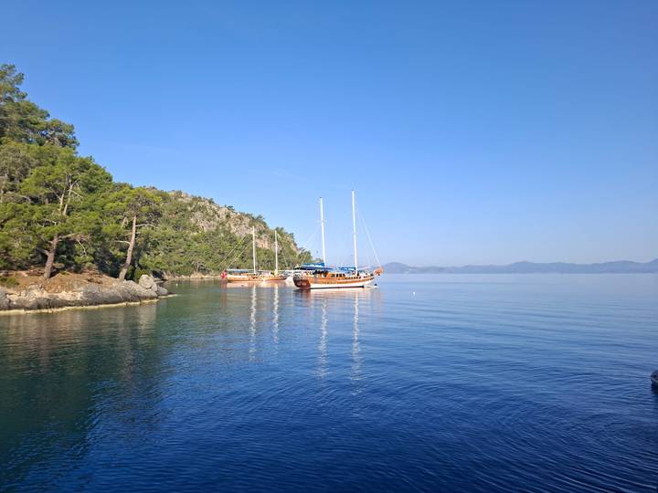 Traditional wooden yachts anchor in a calm turquoise inlet bordered by pine-covered cliffs.