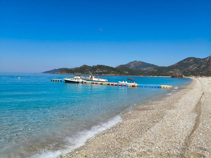 Crystal-clear turquoise sea laps a pebbly beach with moored speedboats and distant hills.