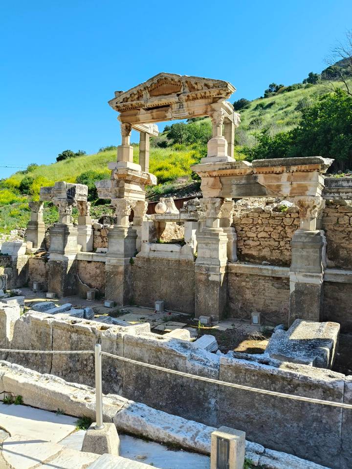 Weathered stone columns and carved capitals of an ancient fountain surrounded by greenery.
