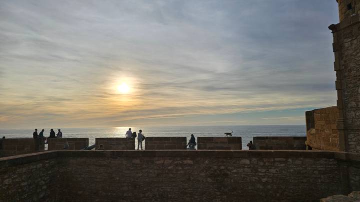 Silhouetted visitors and a dog on seaside ramparts watching the sun set over the Atlantic.