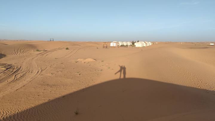 Remote desert camp of white tents in rolling dunes under a pale morning sky with photographer’s shadow.