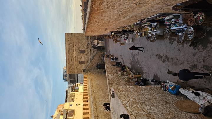 Market alley along fortified stone walls with stalls selling baskets and goods; locals and tourists strolling.