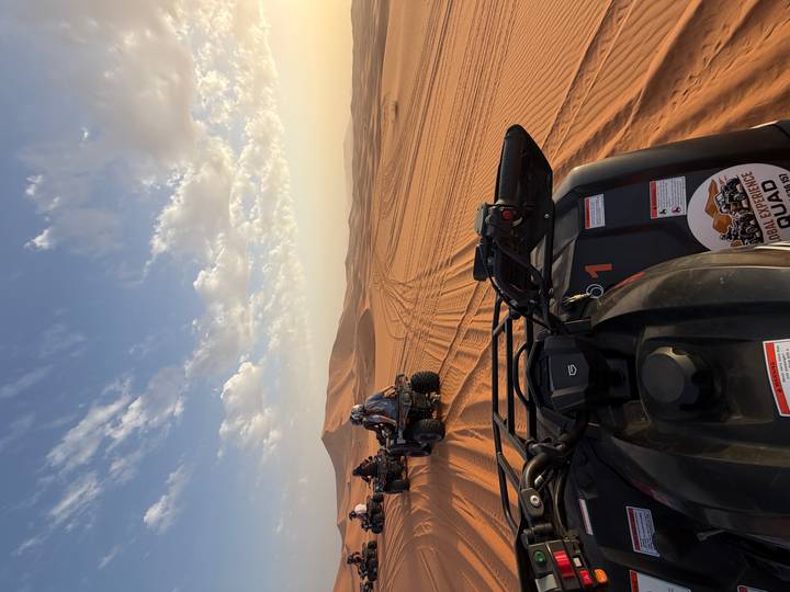 Several riders drive quad bikes across vast orange sand dunes under a blue sky dotted with thin clouds.