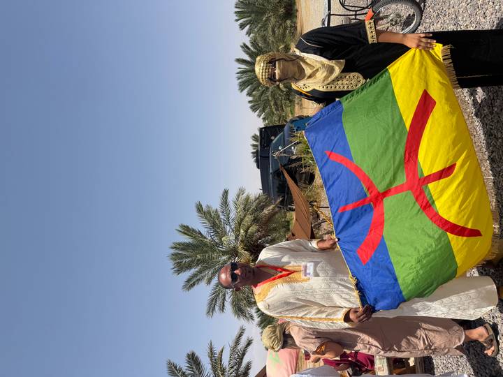 Two people in traditional Moroccan dress proudly hold a colorful Amazigh flag beside date palms on a sunny day.