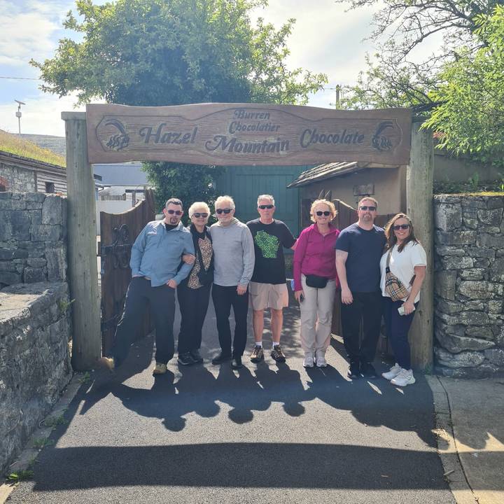 Visitors smiling beneath the wooden sign for Hazel Mountain Chocolate outside a rustic stone entrance.