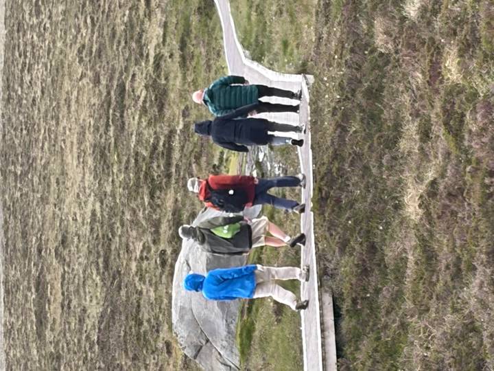 Hikers bundled in jackets walk along a wooden boardwalk across open heathland.