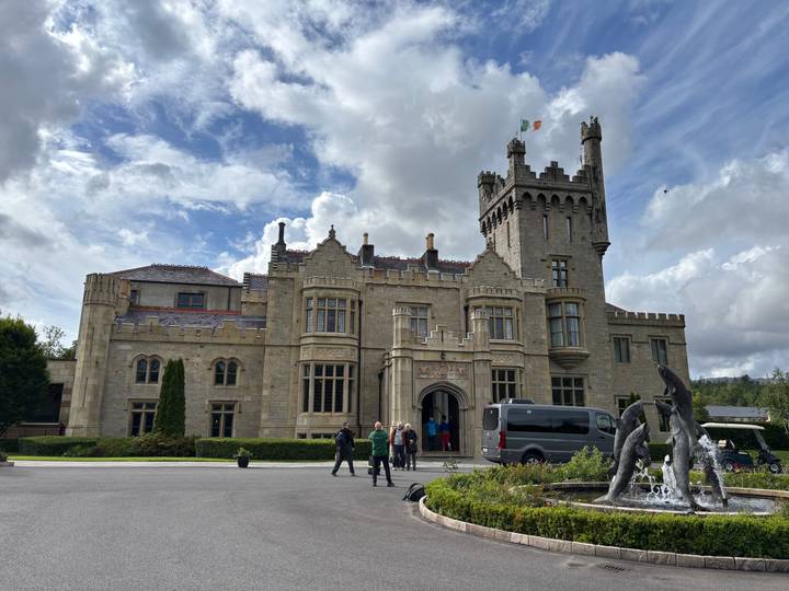 Elegant castle hotel with Irish flag under a dramatic cloudy sky; travellers and a van at the entrance fountain.