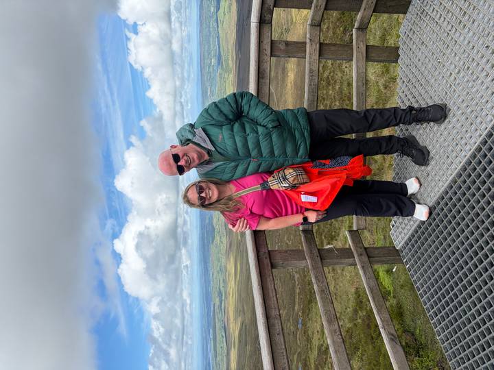 Smiling couple on a wooden viewing platform high above rolling Irish countryside under scattered clouds.