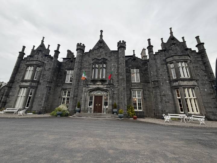 Grey stone Gothic-style manor with Irish and Portuguese flags, white benches and cloudy sky.