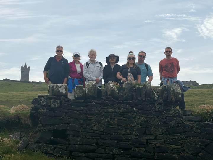 Travellers pose on an old stone wall with a small tower rising on the grassy hill behind.