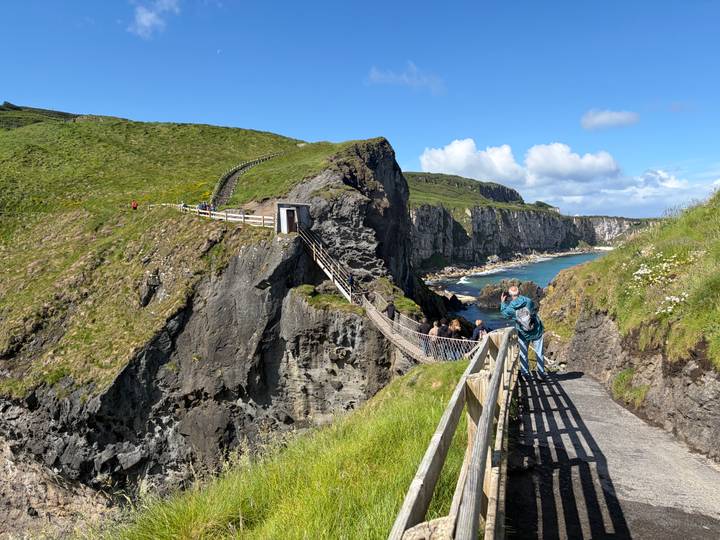 Wooden walkway leads to the dramatic Carrick-a-Rede rope bridge spanning a sea chasm on a sunny day.