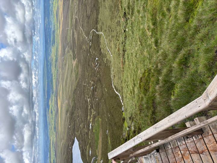 Wide open moorland and rolling hills stretch to the horizon under patchy clouds, seen from a wooden railing viewpoint.