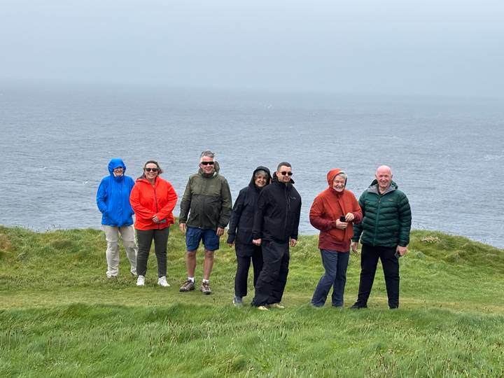Travellers in bright rain jackets stand on wind-swept green cliffs overlooking a grey Atlantic.