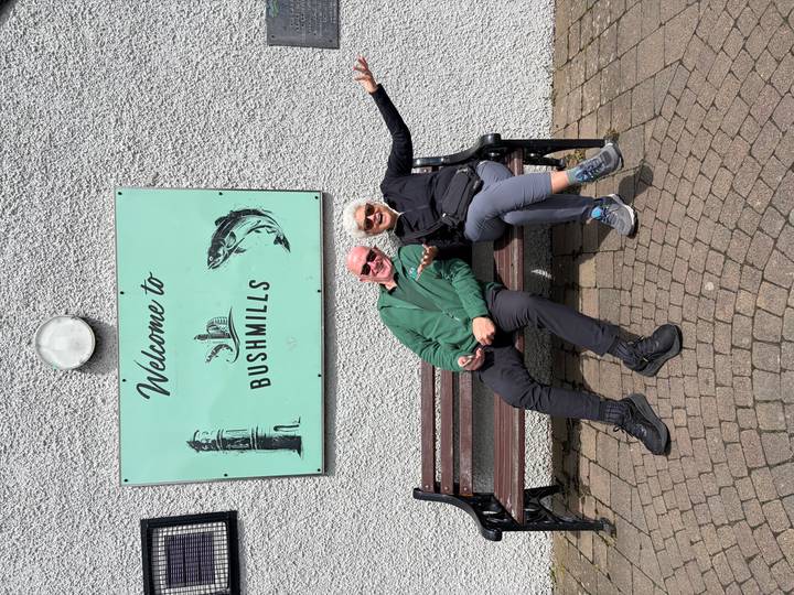 Cheerful couple reclines on a bench beneath a mint-green "Welcome to Bushmills" sign.
