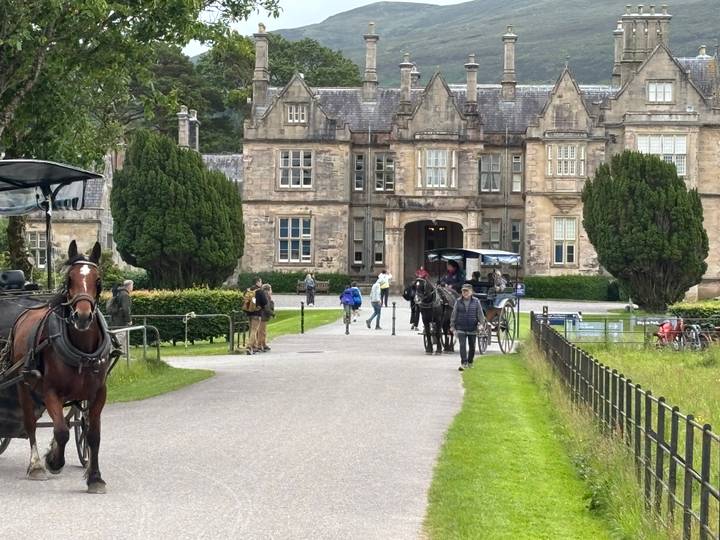 Horse-drawn jaunting cars and visitors approach the stately Muckross House framed by gardens.