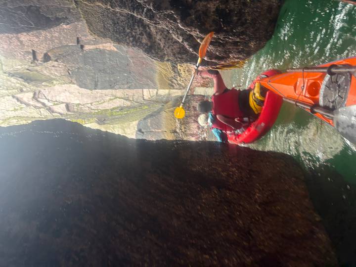 Kayakers paddle through a narrow sea cave corridor between towering rock walls.