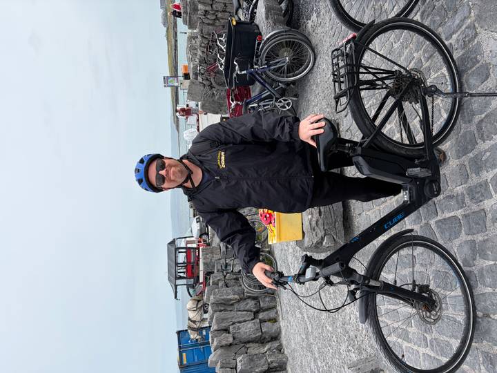 Cyclist wearing helmet poses with an e-bike near stone walls and horse carts by the sea.