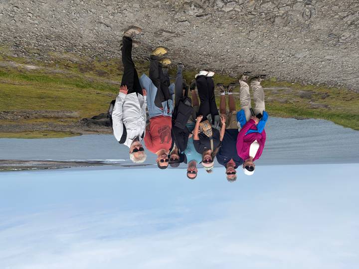 Group of travellers stands on rocky coastal path with sweeping sea views under light blue sky.