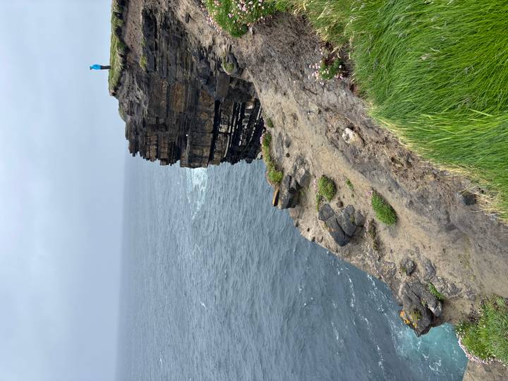 Tall cliff edge with layered rock faces meets choppy grey sea; lone person in blue jacket stands atop.