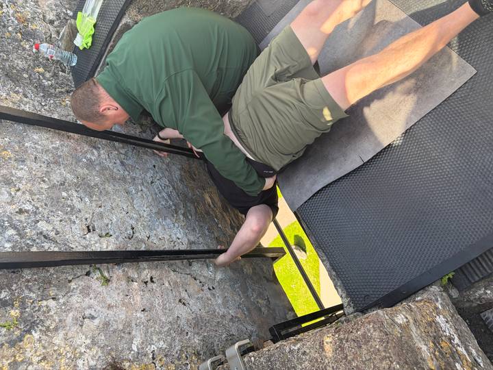 Visitor bends backward to kiss the Blarney Stone while a helper supports him on the castle parapet.