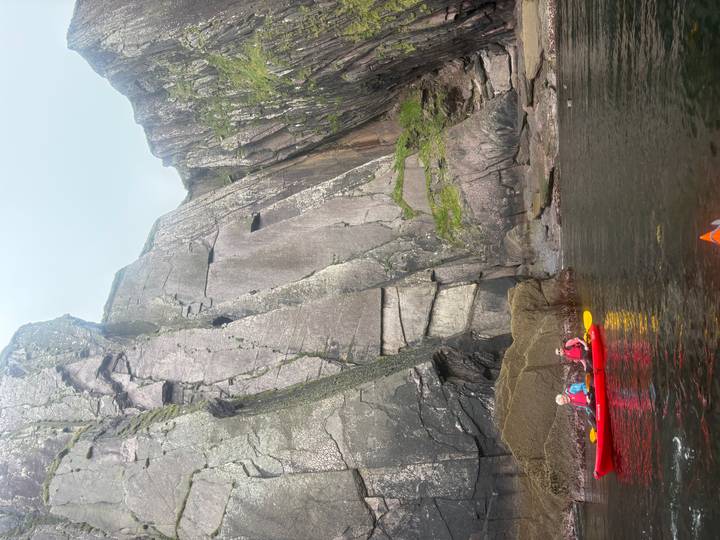Red kayaks float on dark coastal waters beneath towering striated rock cliffs.