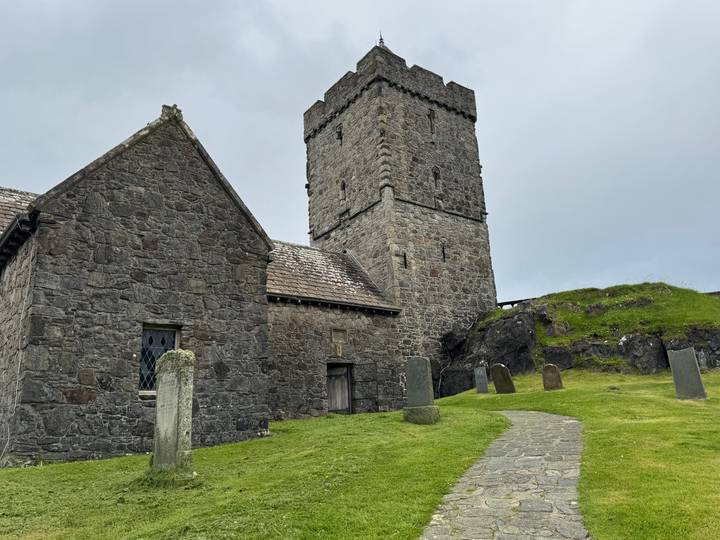 A grey-stone medieval church and tower rise above old gravestones on a grassy knoll.