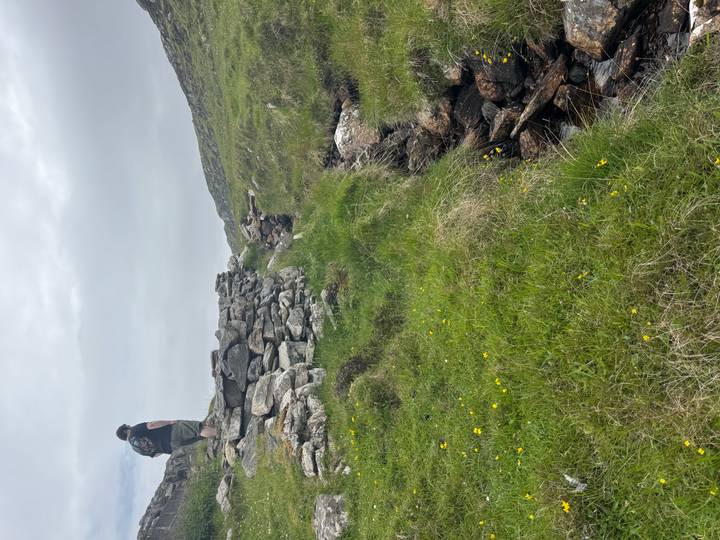 A hiker with backpack stands atop a grassy stone wall overlooking rugged Scottish hills.