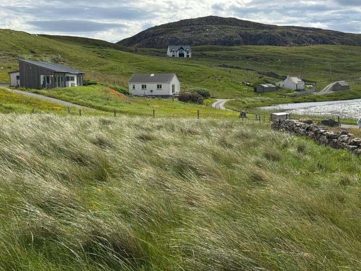 Scattered rural cottages and a tiny chapel sit amid rolling green pasture near a windy coastline.
