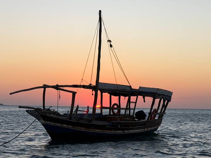 Silhouette of a traditional dhow anchored on calm sea water as the sun sets on the horizon.
