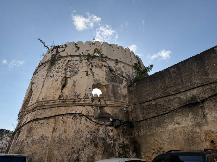 Weathered round tower of an old fort rises against a clear blue sky with greenery sprouting from cracks.