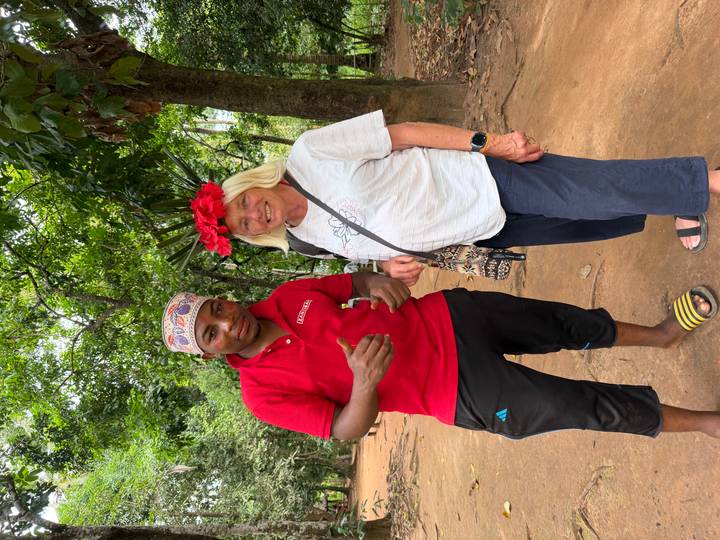 Tourist and local guide smile together in a lush spice farm, one wearing a crown of red flowers.