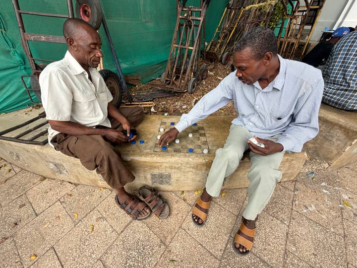 Two men sit opposite each other outdoors playing a board game made of bottle caps on a stone bench.