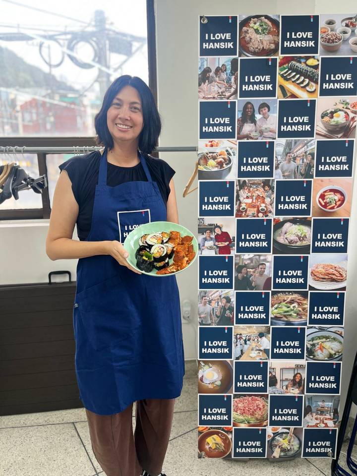 Participant in an apron presenting a plate of homemade kimbap and side dishes beside a poster reading ‘I LOVE HANSIK’.