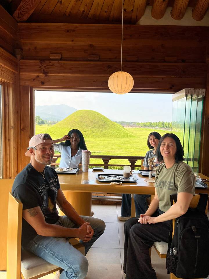 Four friends enjoy a meal at a wooden table with a grassy burial mound and mountains visible through open windows.