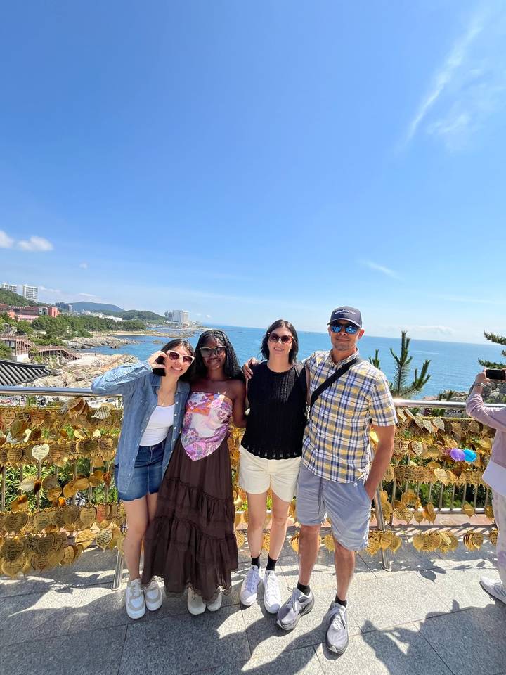 Four friends smile at a coastal viewpoint decorated with golden love locks, azure sea and blue sky behind.