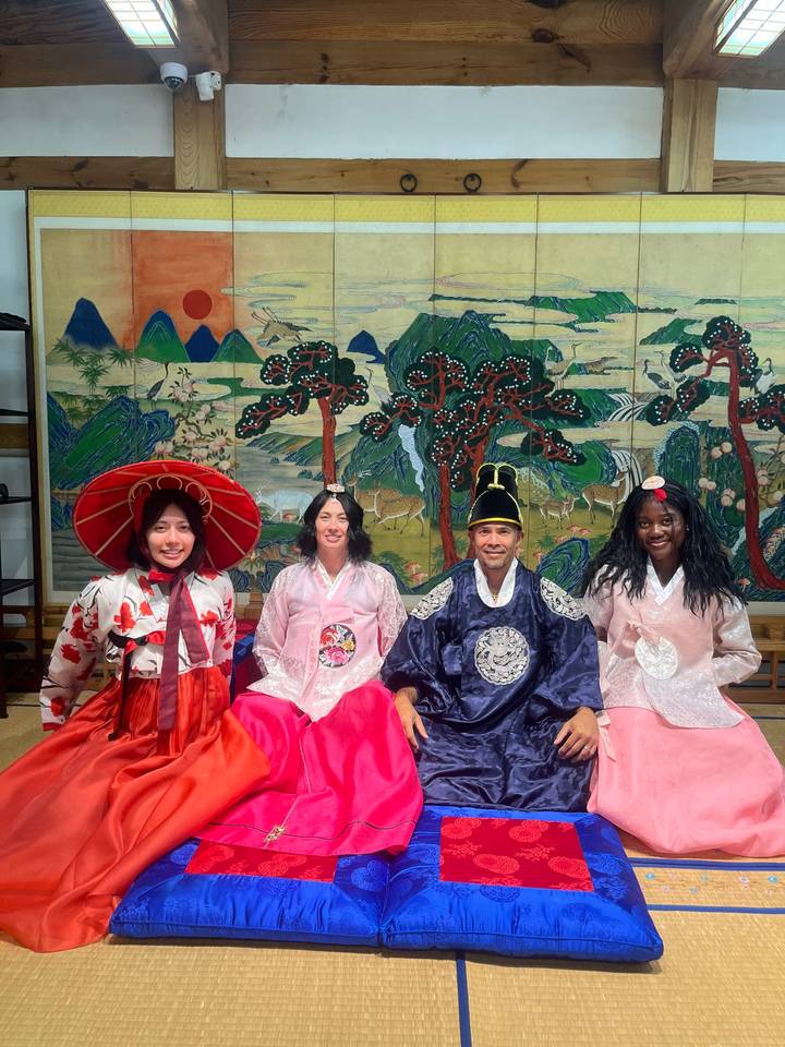 Travelers dressed in traditional hanbok pose in front of a colourful Korean painted screen with deer and pines.